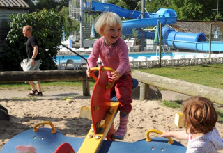 Børn leger på legeplads med vipper ved Vestbirk Camping, med swimmingpool og vandrutsjebane i baggrunden.