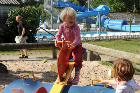 Niños juegan en un balancín en el parque infantil de Vestbirk Camping, con piscina y tobogán al fondo.