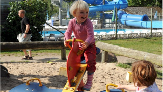 Niños juegan en un balancín en el parque infantil de Vestbirk Camping, con piscina y tobogán al fondo.