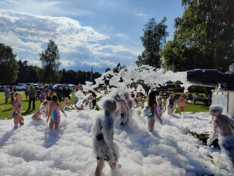 Niños juegan en espuma durante un evento en Vestbirk Camping entre adultos, sol y árboles verdes.
