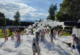 Des enfants jouent dans la mousse lors d’un événement à Vestbirk Camping entourés d’adultes et de verdure.