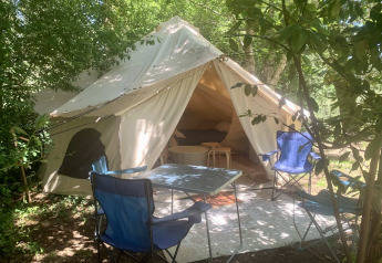 Tienda safari Laurelia XL Family Tent con entrada abierta, sillas al aire libre y mesa bajo los árboles.