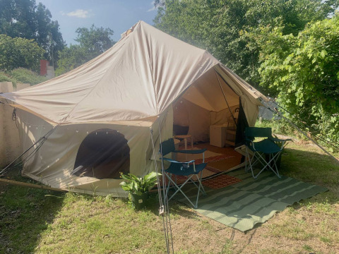 Tienda safari Camelia XL Bell Tent con entrada abierta, dos sillas y alfombra verde, rodeada de naturaleza.