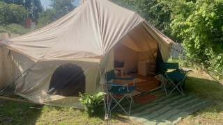Camelia XL Bell Tent safari telt med åbning, to stole og grøn måtte udenfor, omgivet af træer og planter.