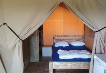 Vista interior de una tienda safari en Camping de Oude Rijn, Países Bajos, con cama, estantería y nevera pequeña.