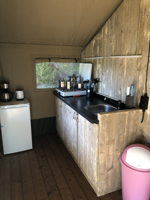 Küchenbereich im Safarizelt mit Holztresen, Spüle, Herd, Kühlschrank und rosa Mülleimer.