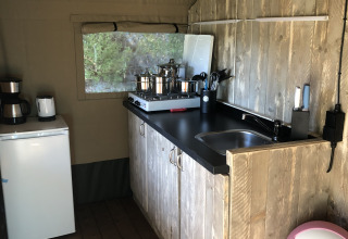 Küchenbereich im Safarizelt mit Holztresen, Spüle, Herd, Kühlschrank und rosa Mülleimer.