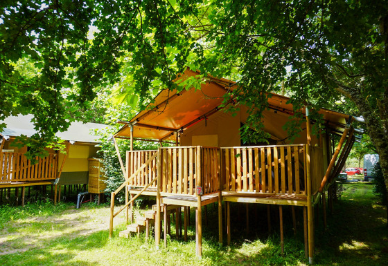 Tienda safari en Camping Pittoresque, Francia, con terraza de madera bajo árboles verdes y frondosos.