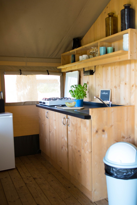 Interior view of a safari tent kitchen with wooden cabinets, stove, and shelves at Camping de Boomgaard, Belgium.