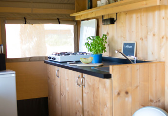 Interior view of a safari tent kitchen with wooden cabinets, stove, and shelves at Camping de Boomgaard, Belgium.