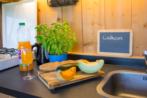 Piano cucina con pane, frutta, succo e erbe accanto a un cartello di benvenuto in una tenda safari in Belgio.