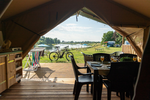 Vista desde dentro de una tienda safari hacia una terraza de madera con mesa, bicicleta y lago al fondo.