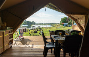 Vista desde dentro de una tienda safari hacia una terraza de madera con mesa, bicicleta y lago al fondo.