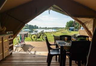 Zicht van binnenuit een safaritent op een houten terras met tafel, fiets en een meer in de verte.