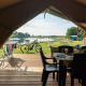 Blick aus dem Inneren eines Safarizelts auf eine Holzterrasse mit Tisch, Fahrrad und See im Hintergrund.