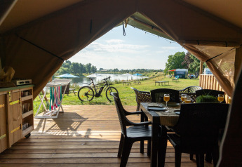 Udsigt fra et safaritelts indre ud over en træterrasse med spisebord, cykel og en sø i baggrunden.