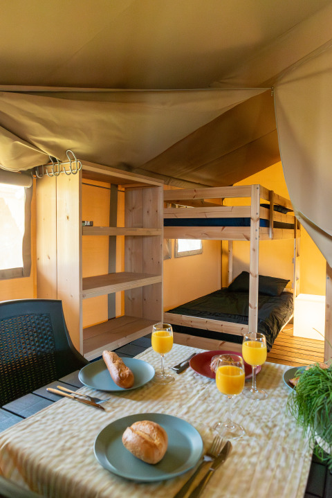 Interieur van een safaritent met stapelbedden en ontbijttafel met broodjes en glazen fruitsap.