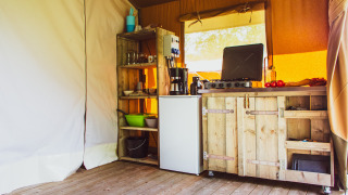 Cozy wooden kitchen setup with stove, fridge and shelves inside a safari tent at Camping de Boomgaard, Belgium.