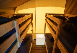Interior de una tienda safari con dos literas de madera, ropa de cama oscura y luz cálida exterior.