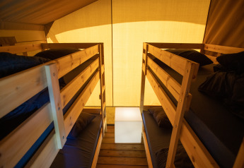 Interno di una tenda safari con due letti a castello in legno, biancheria scura e luce calda.