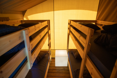 Interior de una tienda safari con dos literas de madera, ropa de cama oscura y luz cálida exterior.