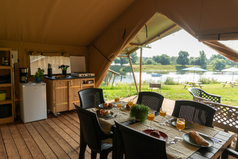 Interno di tenda safari con tavolo da pranzo, cucina e vista fiume al Camping de Boomgaard in Belgio.