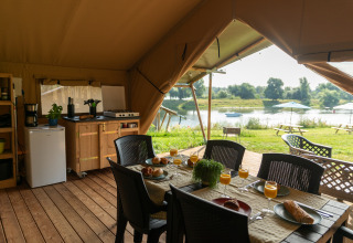 Binnenkant van safaritent met eetplek, keukentje en zicht op de rivier bij Camping de Boomgaard in België.