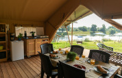 Innenraum eines Safarizelts mit Esstisch, Küche und Blick auf den Fluss bei Camping de Boomgaard in Belgien.