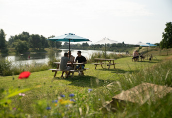 Persone rilassate sotto ombrelloni ai tavoli da picnic vicino al fiume a Safari tent premium Maaszicht, Belgio.