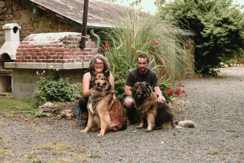 A couple kneels outdoors with two large dogs in a garden at Feather Down Les Jardins des Coccinelles, Brittany.