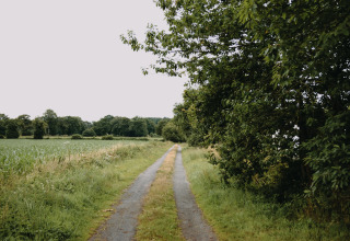 Rural pathway surrounded by green fields and trees near Val-Couesnon, Brittany, France, under a cloudy sky.