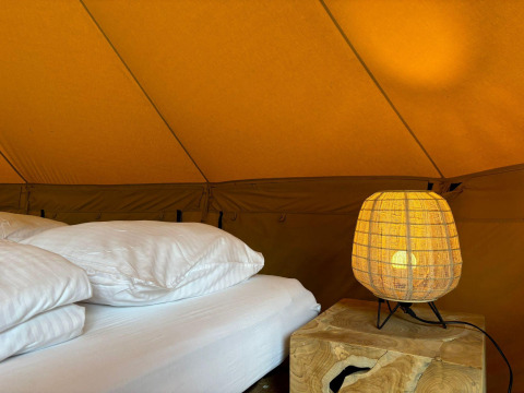 Interior de una tienda safari Bivak Deluxe en Urban-Gardens, Bélgica, con cama, almohadas y lámpara encendida.