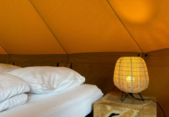 Interior de una tienda safari Bivak Deluxe en Urban-Gardens, Bélgica, con cama, almohadas y lámpara encendida.