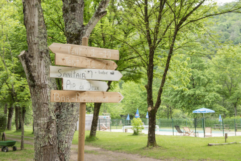 Señales de madera en Camping Pittoresque, Occitania, Francia, rodeadas de árboles y área de piscina.