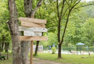 Wooden directional signs at Camping Pittoresque in Occitanie, France, surrounded by trees and lounge chairs.