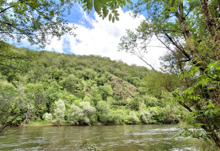 River with lush green trees and hills at Camping Pittoresque in Occitanie, France, under a partly cloudy sky.