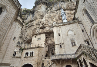 Historic stone buildings blend into a cliffside at Camping Pittoresque, Occitanie, France, under cloudy skies.