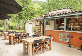 Terraza exterior de Camping Pittoresque, Occitanie, Francia, con mesas, sillas de madera y barra de bar.