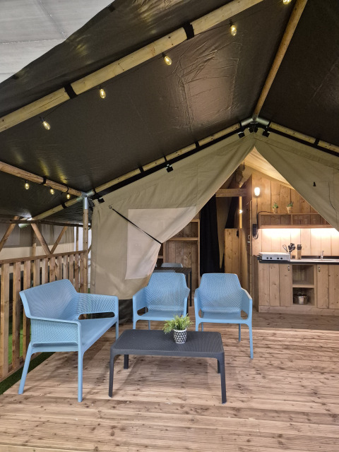 Interior of a safari tent with blue chairs, coffee table, wooden deck, kitchen area, and warm lighting.