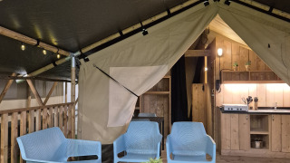 Interior of a safari tent with blue chairs, coffee table, wooden deck, kitchen area, and warm lighting.