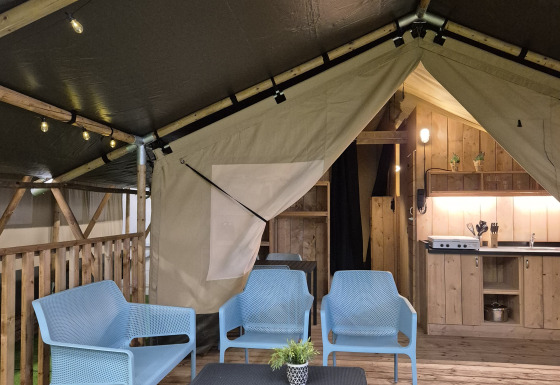Interior of a safari tent with blue chairs, coffee table, wooden deck, kitchen area, and warm lighting.