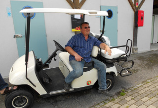 Mand sidder smilende i en golfvogn foran Camping Seasonova Les Marguerites i Hauts-de-France, Frankrig.