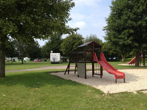 Parque infantil con tobogán rojo y arena, rodeado de árboles y autocaravanas en Camping Seasonova Les Marguerites.