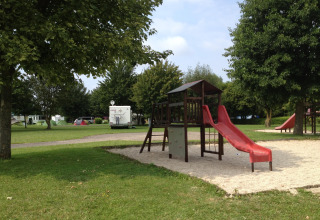 Aire de jeux avec toboggan rouge sur sable, entourée d’arbres et de camping-cars au Camping Seasonova Les Marguerites.