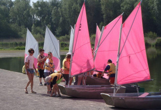 Børn og voksne forbereder små sejlbåde med lyserøde og hvide sejl ved søen i Camping Seasonova Les Marguerites.