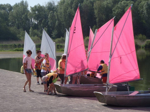 Kinder und Erwachsene bereiten kleine Segelboote mit pinken und weißen Segeln am See bei Camping Seasonova Les Marguerites vor.