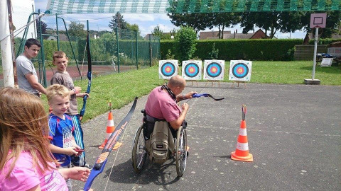 Persona in sedia a rotelle fa tiro con l’arco con bambini all’aperto in un campeggio di Hauts-de-France.
