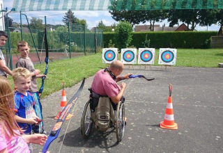 Persona in sedia a rotelle fa tiro con l’arco con bambini all’aperto in un campeggio di Hauts-de-France.