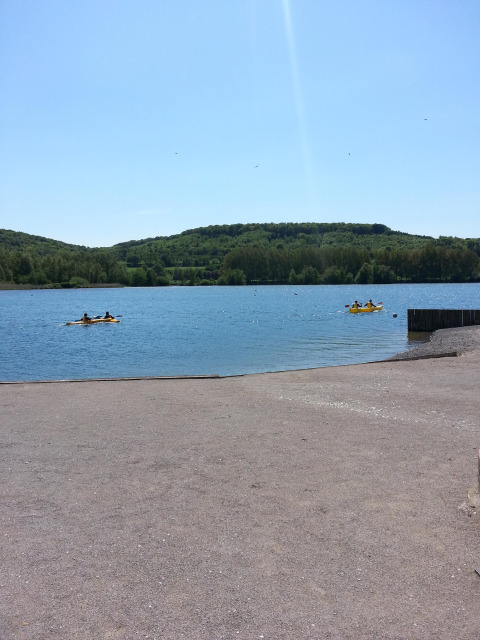 Vista di un lago con colline sullo sfondo e persone su pedalò gialli al Camping Seasonova Les Marguerites.