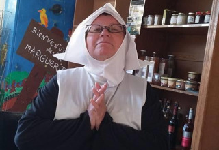 Eine als Nonne verkleidete Person sitzt mit gefalteten Händen vor einem Regal im Camping Seasonova Les Marguerites, Frankreich.